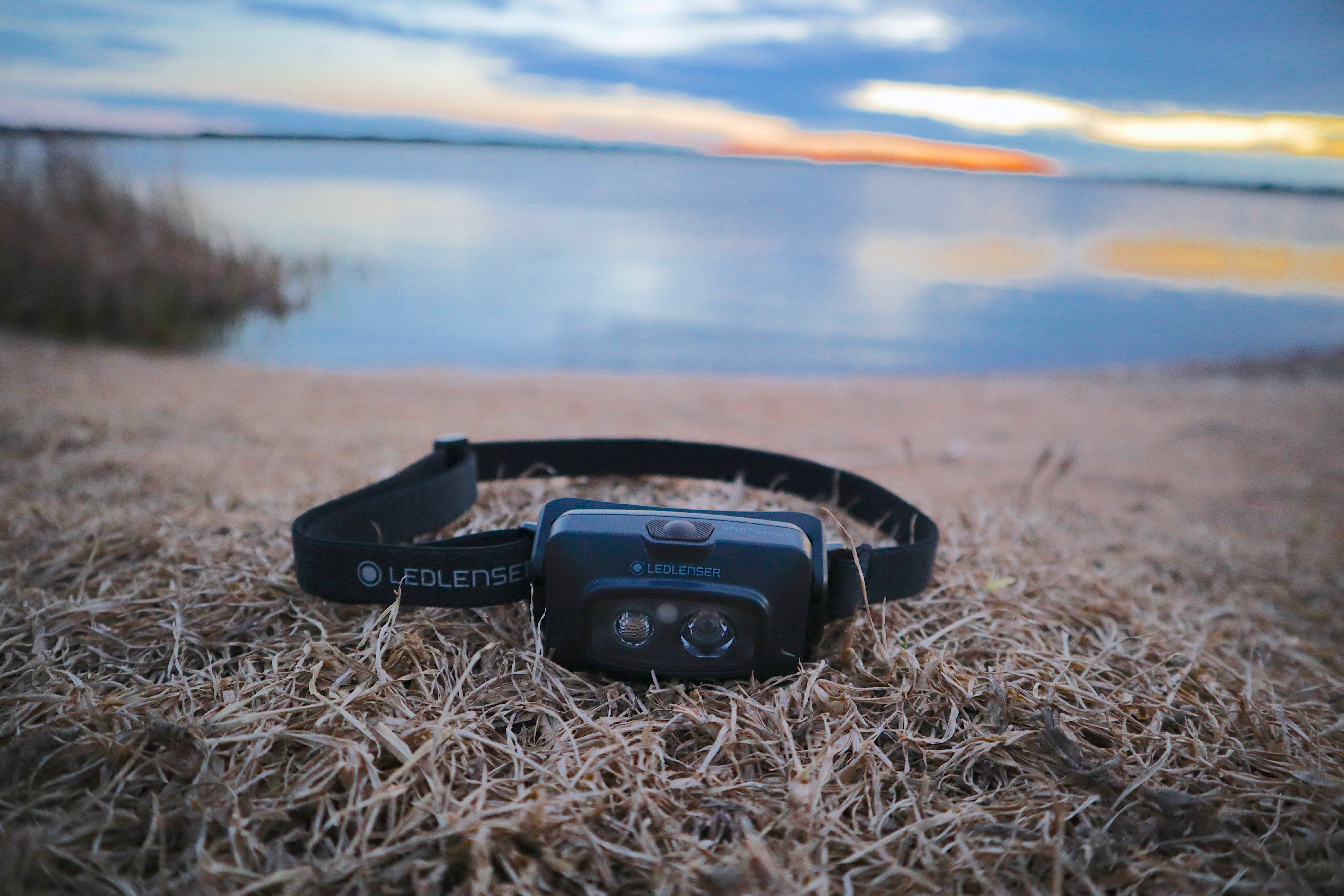 Ledlenser HF4R Signature headlamp resting on dry grass at a lakeside campsite at dusk, unlit