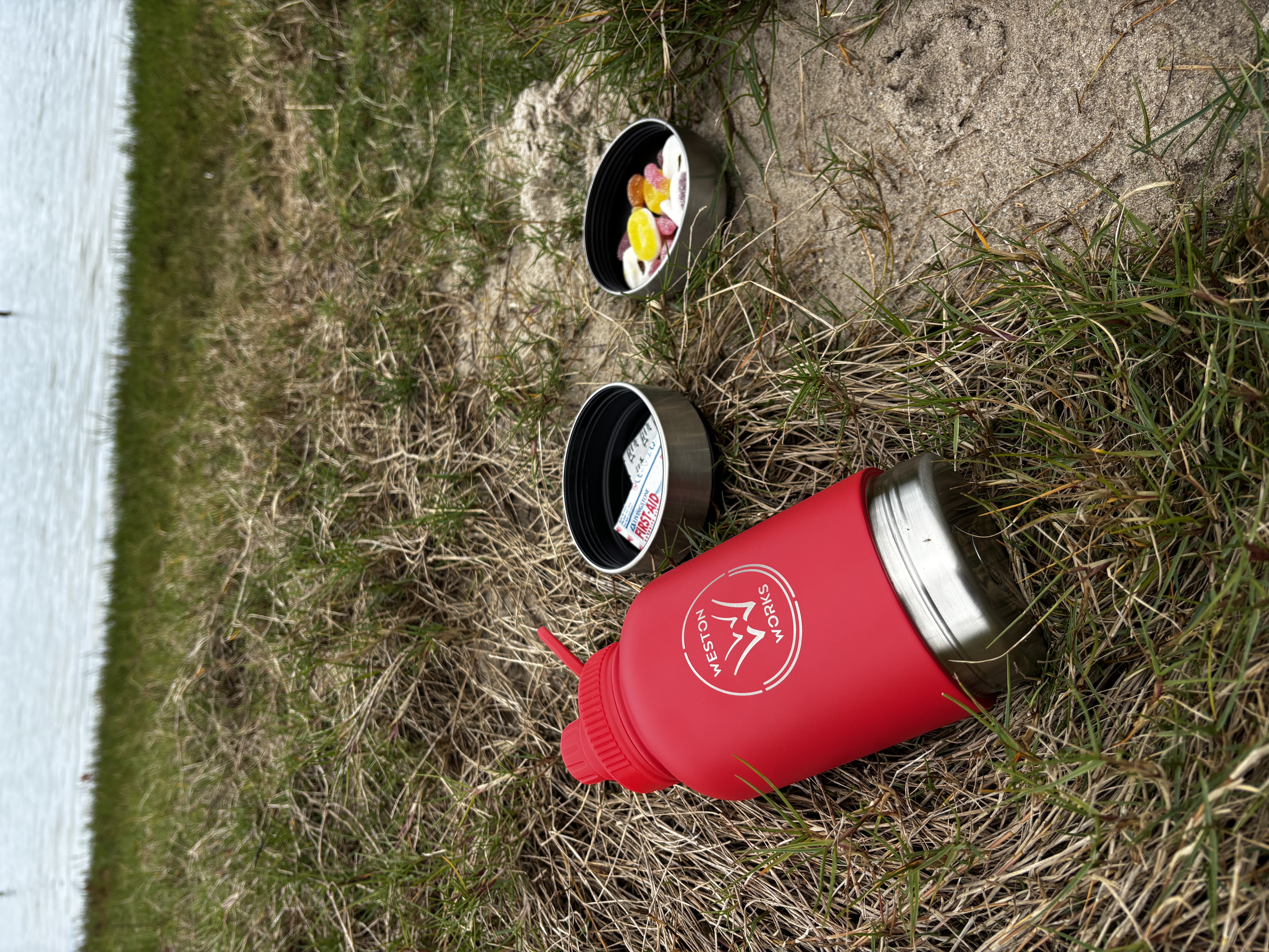WestonWorks First Aid Bottle lying in grass with both storage cups open, one holding bandages and one holding lollies